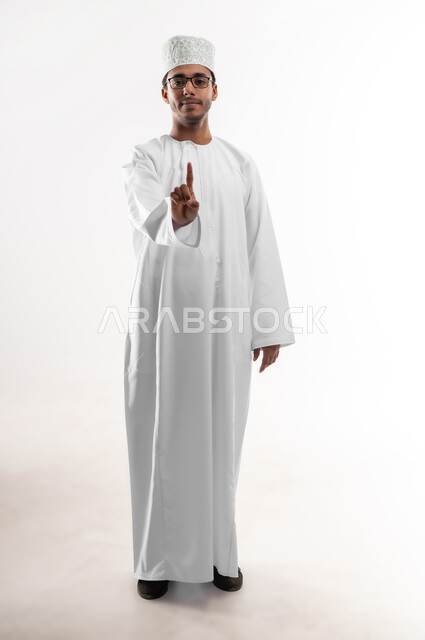 الاهتمام بالأناقة والمظهر الخارجي، تعابير السرور والثقة بالنفس، بورتريه لشاب عربي خليجي عماني يرتدي الدشداشة و الكمه والنظارات الطبية ينظر الى الكاميرا يشير بإصبع السبابة بدلالة الرقم واحد، صورة الجسم بالطول الكامل، خلفية بيضاء