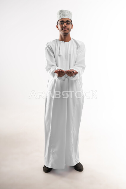 Using advanced technology, displaying mobile applications and services, close-up portrait of a young Omani Gulf Arab man wearing a dishdasha, a turban and glasses looking at the camera holding a mobile phone with a blank black screen, e-shopping concept, full-length body image, white background