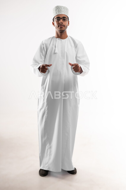 Concept of presenting and displaying a certain item, measuring and estimating the size of things, portrait of a smiling young Arab Gulf Omani man wearing a dishdasha, a sleeve and eyeglasses, looking at the camera, raising his hands up, holding a holographic element, expressions of taking pictures, full-length body image, white background