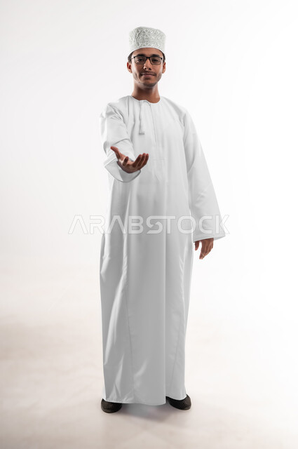 إمساك و إحتواء عنصر ما، مفهوم الاناقة و الاهتمام بالمظهر الخارجي, بورتريه مقرب لشاب عربي خليجي عماني يرتدي الدشداشة و الكمه والنظارات الطبية ينظر إلى الكاميرا يمد يده للأمام يحاول التقاط عناصر هولوجرامية ثلاثية الأبعاد، صورة الجسم بالطول الكامل، خلفية بيضاء
