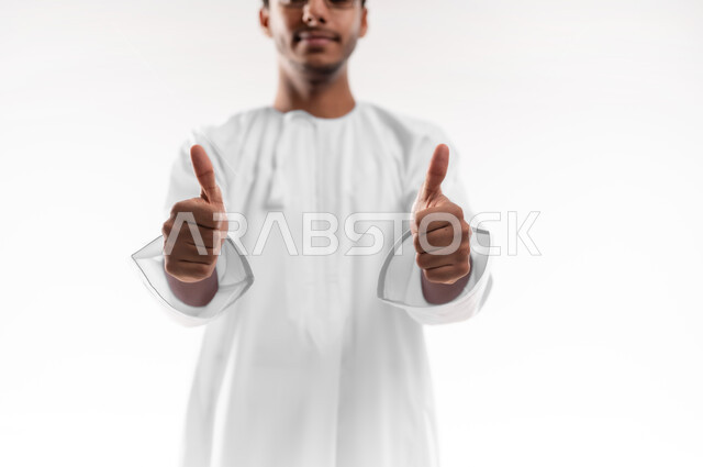 الشعور بالرضا والقبول، مفهوم النجاح وتحقيق الأهداف، بورتريه مقرب لشاب عربي خليجي عماني يرتدي الدشداشة يرفع اصبع الابهام للاعلى بايماءات الجودة والامتياز، تعابير الفرح والسرور، خلفية بيضاء