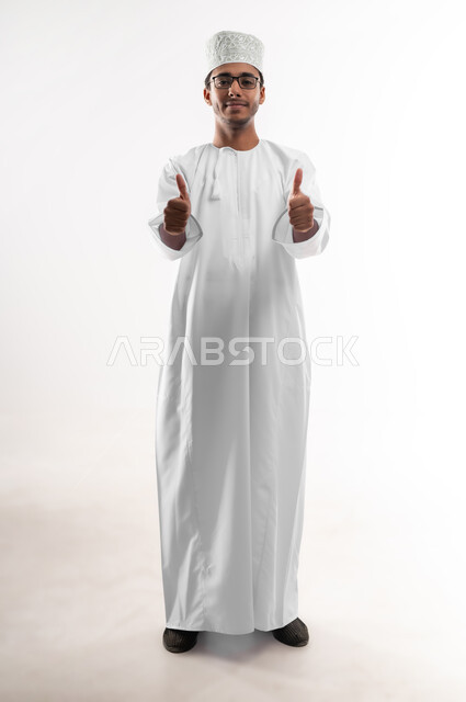 الشعور بالرضا والقبول، مفهوم النجاح وتحقيق الأهداف، بورتريه لشاب عربي خليجي عماني يرتدي الدشداشة والكمه والنظارات الطبية يرفع اصبع الابهام للاعلى بايماءات الجودة والامتياز، تعابير الفرح والسرور، صورة الجسم بالطول الكامل, خلفية بيضاء