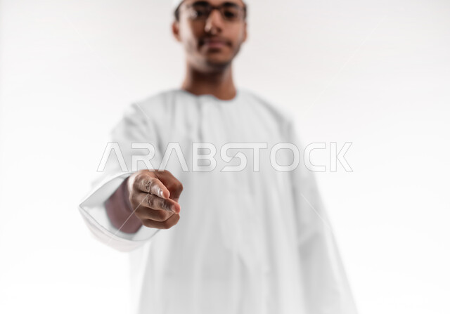 Concept of clicking, pressing and interacting with holographic elements, determining a specific location, close-up portrait of a young Arab Gulf Omani man wearing a dishdasha, a sleeve and glasses looking at the camera, raising his hand forward, pointing with his index finger to a specific target, white background