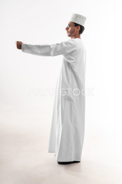 الترحيب و السلام و إلقاء التحية، بناء علاقات و تكوين صداقات جديدة، بورتريه لشاب عربي خليجي عماني يرتدي الدشداشة والكمه والنظارة الطبية يقوم بتوجيه قبضة يده بإيماءات القوة والإصرار والمواجهة، صورة الجسم بالطول الكامل، خلفية بيضاء
