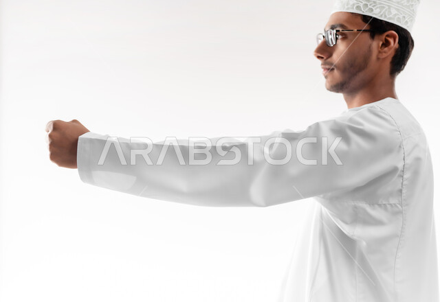 السلام و الترحيب و إلقاء التحية، بناء علاقات و تكوين صداقات جديدة، بورتريه مقرب لشاب عربي خليجي عماني يرتدي الدشداشة والكمه والنظارة الطبية يقوم بتوجيه قبضة يده بإيماءات القوة والإصرار والمواجهة، خلفية بيضاء