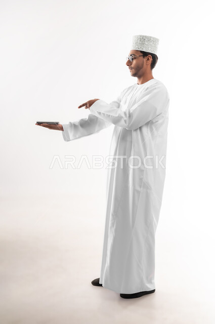 استخدام التكنولوجيا المتطورة، عرض تطبيقات وخدمات الهاتف المحمول، بورتريه لشاب عربي خليجي عماني يرتدي الدشداشة و الكمه والنظارات الطبية يحمل بيده جوال بشاشة سوداء فارغة ويشير اليه باصبع السبابة، صورة الجسم بالطول الكامل، خلفية بيضاء