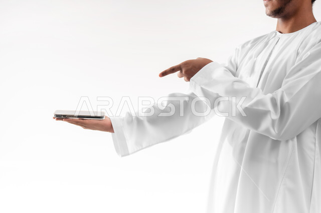 Using advanced technology, displaying mobile applications and services, close-up portrait of a young Gulf Arab Omani man wearing a dishdasha, a sleeve and glasses, holding a mobile phone with a blank black screen and pointing at it with his index finger, white background