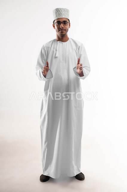 Trying to estimate the size of objects, holding and containing an item, close-up portrait of a young Arab Gulf Omani man wearing a dishdasha, a sleeve and glasses looking at the camera, opening his hands to display 3D holographic elements, full-length body image, white background