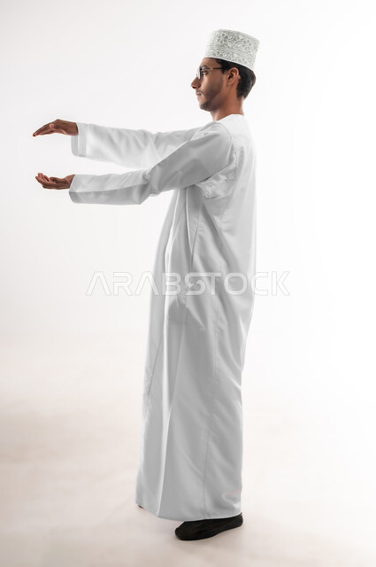 إحتواء وإمساك عنصر ما، مفهوم الاناقة و الاهتمام بالمظهر الخارجي, بورتريه لشاب عربي خليجي عماني يرتدي الدشداشة و الكمه والنظارات الطبية يفتح يديه لعرض عناصر هولوجرامية ثلاثية الأبعاد، صورة الجسم بالطول الكامل, خلفية بيضاء
