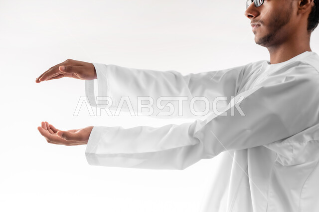 إمساك وإحتواء عنصر ما، مفهوم الاناقة و الاهتمام بالمظهر الخارجي, بورتريه مقرب لشاب عربي خليجي عماني يرتدي الدشداشة و الكمه والنظارات الطبية يفتح يديه لعرض عناصر هولوجرامية ثلاثية الأبعاد، خلفية بيضاء