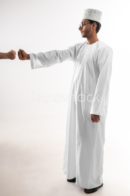 بناء علاقات و تكوين صداقات جديدة، الترحيب و السلام و إلقاء التحية، بورتريه لشاب عربي خليجي عماني يرتدي الدشداشة والكمه والنظارة الطبية يقوم بتوجيه قبضة يده مقابل يد شخص آخر، ايماءات وحركات يدين تدل على التشجيع، صورة الجسم بالطول الكامل، خلفية بيضاء
