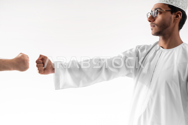 بناء علاقات و تكوين صداقات جديدة، الترحيب و السلام و إلقاء التحية، بورتريه مقرب لشاب عربي خليجي عماني يرتدي الدشداشة والكمه والنظارة الطبية يقوم بتوجيه قبضة يده مقابل يد شخص آخر، ايماءات وحركات يدين تدل على التشجيع، خلفية بيضاء