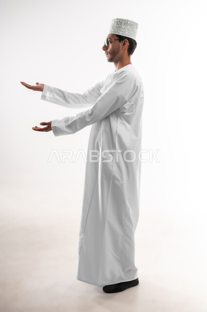 رفع وحمل شيء ما، استخدام حاسة اللمس لتحريك العناصر الهولوجراميه، بورتريه لشاب عربي خليجي عماني يرتدي الدشداشة والنظارات الطبية يرفع يده للاعلى بإيماءات الموازنة وتقدير الحجم، صورة الجسم بالطول الكامل، خلفية بيضاء