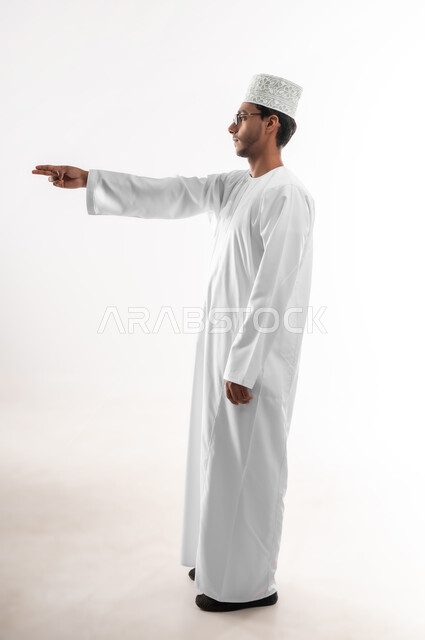 مفهوم الاختيار و تحديد موقع معين، بورتريه لشاب عربي خليجي عماني يرتدي الدشداشة والنظارات الطبية يرفع يده للاعلى يشير الى هدف محدد، حركات يدين تدل على التحذير والتنبيه، صورة الجسم بالطول الكامل، خلفية بيضاء