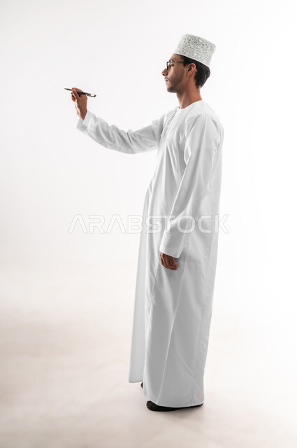 تدوين وتسجيل الملاحظات، الإستعداد للكتابة وتحديد شيء ما, بورتريه لشاب عربي خليجي عماني يرتدي الدشداشة والكمه والنظارات الطبية يمسك بيده قلم يكتب شيء ما, مفهوم التعليم بالمناهج المقررة، صورة الجسم بالطول الكامل, خلفية بيضاء