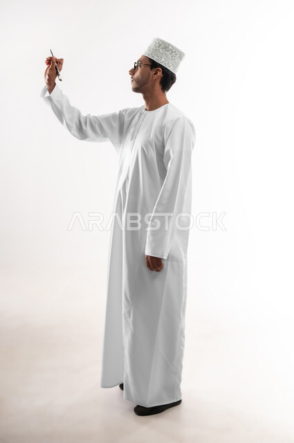 تدوين وتسجيل الملاحظات، الإستعداد للكتابة وتحديد شيء ما, بورتريه لشاب عربي خليجي عماني يرتدي الدشداشة والكمه والنظارات الطبية يمسك بيده قلم يكتب شيء ما, مفهوم التعليم بالمناهج المقررة، صورة الجسم بالطول الكامل, خلفية بيضاء
