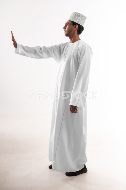 مفهوم الرفض والامتناع والانزعاج, ايماءات الحزم والجدية, طلب التوقف وعدم الاقتراب, بورتريه لشاب عربي خليجي عماني يرتدي الدشداشة والكمه والنظارات الطبية يمد يده نحو الأمام مع رفع الكف بتعابير النهي عن فعل امر ما, صورة الجسم بالطول الكامل, خلفية بيضاء