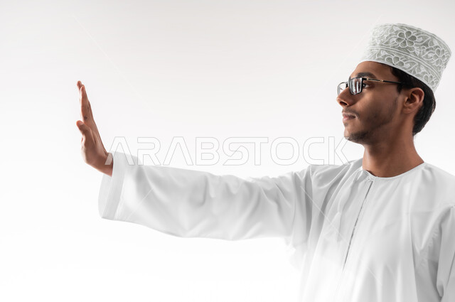 مفهوم الرفض والامتناع والانزعاج, ايماءات الحزم والجدية, طلب التوقف وعدم الاقتراب, بورتريه مقرب لشاب عربي خليجي عماني يرتدي الدشداشة والكمه والنظارات الطبية يمد يده نحو الأمام مع رفع الكف بتعابير النهي عن فعل امر ما, خلفية بيضاء