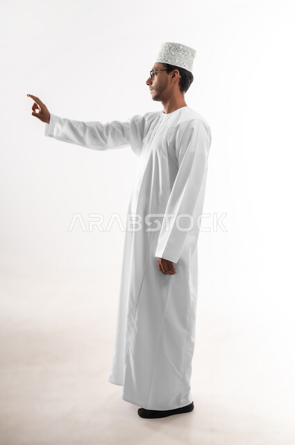 Interaction with holographic elements, concept of selection and positioning, portrait of a young Omani Gulf Arab man wearing a dishdasha, a sleeve and glasses, raising his hand up pointing to a specific target, full-length body image, white background
