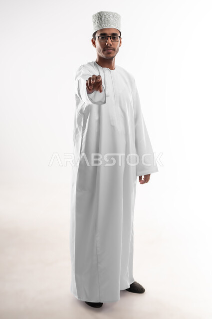 احتواء وإمساك عنصر ما، مفهوم الاناقة و الاهتمام بالمظهر الخارجي, بورتريه لشاب عربي خليجي عماني يرتدي الدشداشة و الكمه والنظارات الطبية ينظر إلى الكاميرا يمد يده للأمام يحاول التقاط عناصر هولوجرامية ثلاثية الأبعاد، صورة الجسم بالطول الكامل، خلفية بيضاء
