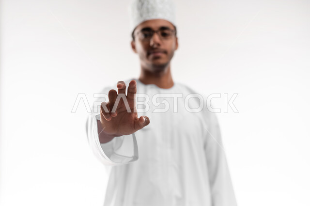 إمساك و إحتواء عنصر ما، مفهوم الاناقة و الاهتمام بالمظهر الخارجي, بورتريه مقرب لشاب عربي خليجي عماني يرتدي الدشداشة و الكمه والنظارات الطبية ينظر إلى الكاميرا يمد يده للأمام يحاول التقاط عناصر هولوجرامية ثلاثية الأبعاد، خلفية بيضاء