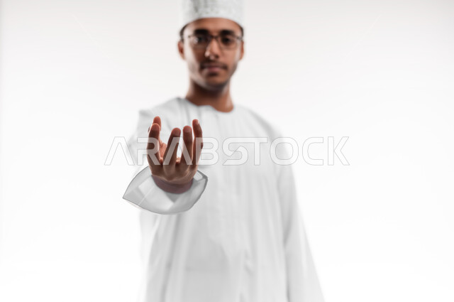 إمساك و إحتواء عنصر ما، مفهوم الاناقة و الاهتمام بالمظهر الخارجي, بورتريه مقرب لشاب عربي خليجي عماني يرتدي الدشداشة و الكمه والنظارات الطبية ينظر إلى الكاميرا يمد يده للأمام يحاول التقاط عناصر هولوجرامية ثلاثية الأبعاد، خلفية بيضاء