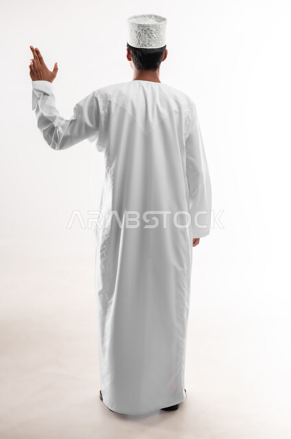 Gestures and expressions of the number three, specifying a specific location, portrait from the back of a young Arab Gulf Omani man wearing a dishdasha, a sleeve and glasses, raising his hand up pointing to a specific target, full-length body image, white background