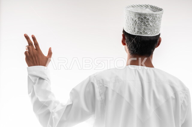 إيماءات و تعابير الرقم ثلاثة، تحديد موقع معين، بورتريه مقرب من الخلف لشاب عربي خليجي عماني يرتدي الدشداشة والكمه والنظارات الطبية يرفع يده للاعلى يشير الى هدف محدد، خلفية بيضاء