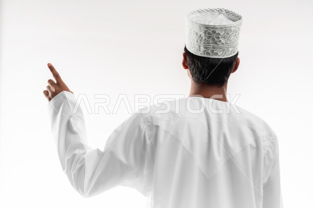 تحديد موقع معين، بورتريه مقرب من الخلف لشاب عربي خليجي عماني يرتدي الدشداشة والكمه والنظارات الطبية يرفع يده للاعلى يشير بأصبع السبابة الى هدف محدد، مفهوم النقر والضغط والتفاعل مع العناصر الهولوجرامية، خلفية بيضاء