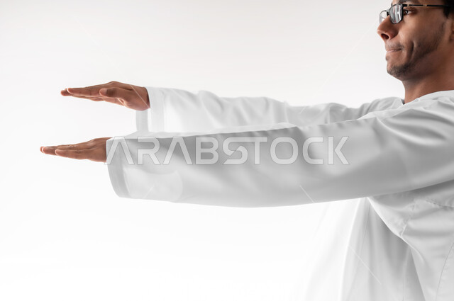 Estimating and measuring the size of objects, the concept of presenting and displaying a certain item, a close-up portrait of a young Gulf Arab Omani man wearing a dishdasha, a sleeve and glasses, raising his hand up holding a holographic element, white background
