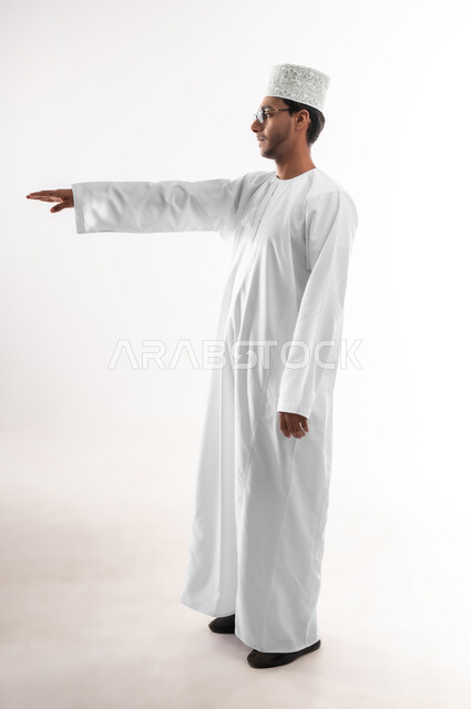 Concept of presenting and displaying a certain item, estimating and measuring the size of things, portrait of a young Arab Gulf Omani man wearing a dishdasha, a sleeve and glasses, raising his hand up trying to touch the holographic elements, full-length body image, white background