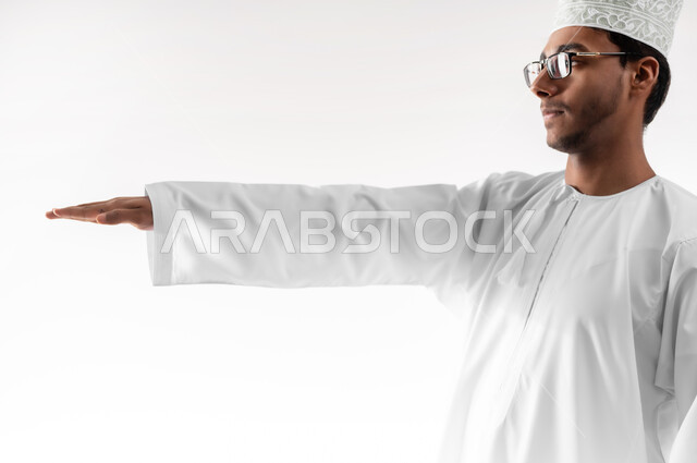 مفهوم تقديم وعرض عنصر معين، تقدير وقياس حجم الأشياء، بورتريه مقرب لشاب عربي خليجي عماني يرتدي الدشداشة والكمه والنظارات الطبية يرفع يده للاعلى يحاول لمس العناصر الهولوجرامية، خلفية بيضاء