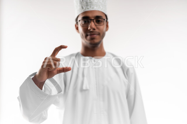 تقدير وقياس حجم الأشياء، مفهوم تقديم وعرض عنصر معين، بورتريه لشاب عربي خليجي عماني يرتدي الدشداشة والكمه والنظارات الطبية ينظر الى الكاميرا يرفع يده للاعلى يمسك عنصر هولوجرامي، خلفية بيضاء