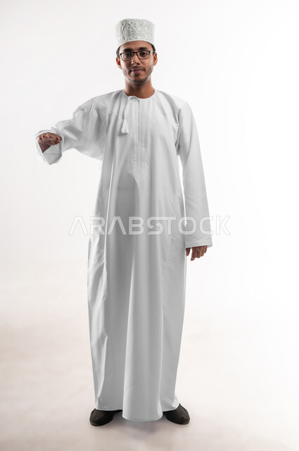 قياس حجم الأشياء، مفهوم تقديم وعرض عنصر معين، بورتريه لشاب عربي خليجي عماني يرتدي الدشداشة والكمه والنظارات الطبية ينظر الى الكاميرا يرفع يده للاعلى بإيماءات تحديد طول شيء ما، صورة الجسم بالطول الكامل, خلفية بيضاء