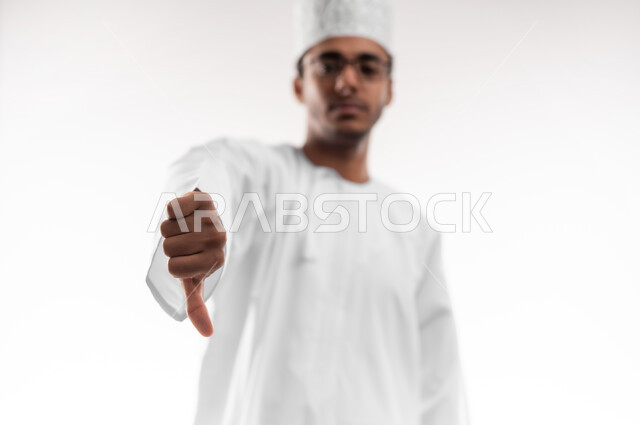 الشعور بالإنزعاج من شيء ما، مفهوم الرفض والفشل، بورتريه مقرب لشاب عربي خليجي عماني يرتدي الدشداشة والكمه والنظارات الطبية يخفض ابهامه بايماءات الرفض وعدم القبول، تعابير التوتر وعد الرضا، خلفية بيضاء
