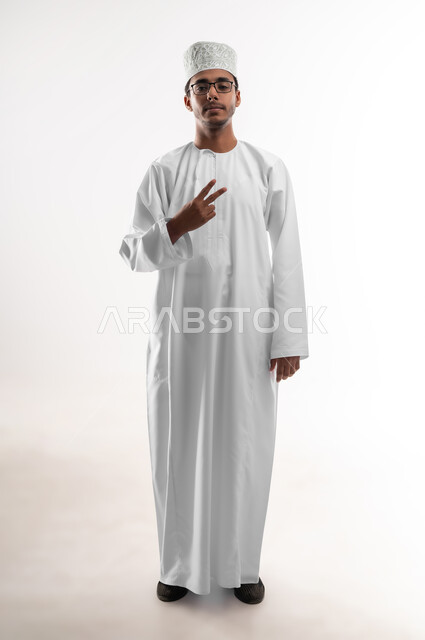 النجاح وتحقيق الأهداف, مفهوم السعادة بالوصول إلى الغايات, بورتريه لشاب عربي خليجي عماني يرتدي الدشداشة والكمه والنظارات الطبية يرفع يده بإشارة النصر والقوة, مفهوم الفوز والانتصار, النظر إلى الكاميرا بتعابير الثقة بالنفس, صورة الجسم بالطول الكامل, خلفية بيضاء