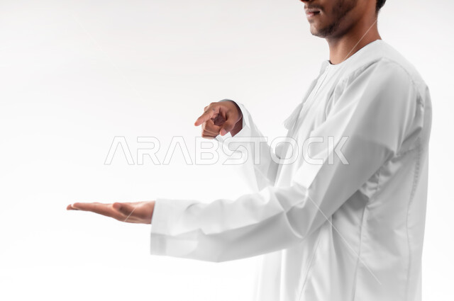 محاولة رفع عناصر هولوجرامية ثلاثية الأبعاد، مفهوم الاناقة و الاهتمام بالمظهر الخارجي, بورتريه مقرب لشاب عربي خليجي عماني يرتدي الدشداشة يشير بإصبع السبابة الى كفه يحمل عنصر ما، خلفية بيضاء