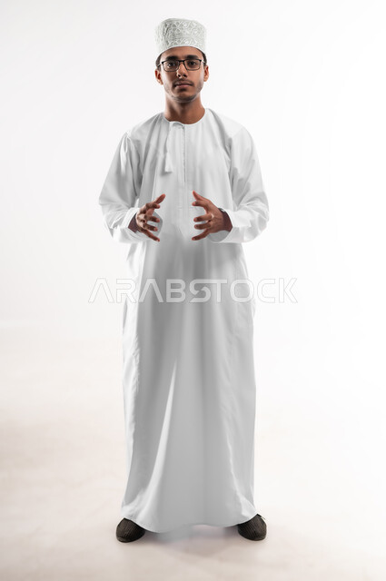إحتواء وإمساك عنصر ما، مفهوم الاناقة و الاهتمام بالمظهر الخارجي, بورتريه لشاب عربي خليجي عماني يرتدي الدشداشة و الكمه والنظارات الطبية ينظر إلى الكاميرا يفتح يديه لعرض عناصر هولوجرامية ثلاثية الأبعاد، صورة الجسم بالطول الكامل, خلفية بيضاء
