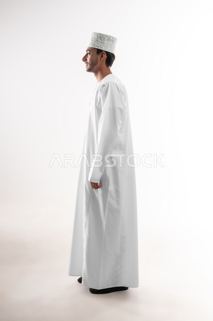 مفهوم الاناقة و الاهتمام بالمظهر الخارجي, بورتريه من الجانب لشاب عربي خليجي عماني يرتدي الدشداشة و الكمه ينظر إلى شيء ما بإيماءات التركيز، صورة الجسم بالطول الكامل, خلفية بيضاء