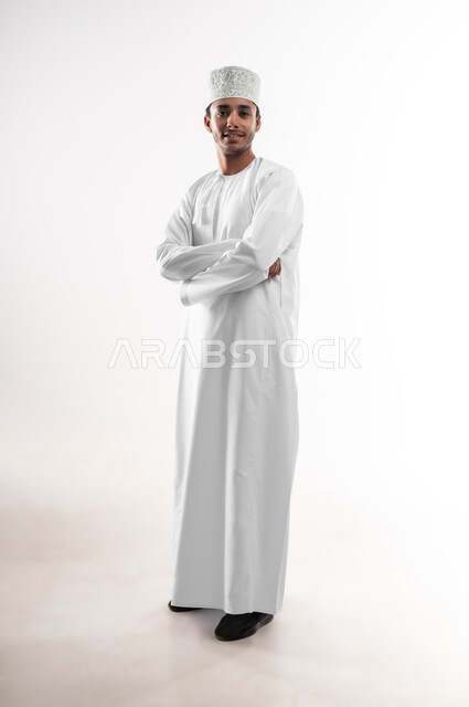 مفهوم الاناقة و الاهتمام بالمظهر الخارجي, بورتريه لشاب عربي خليجي عماني يرتدي الدشداشة و الكمه ينظر إلى الكاميرا بإيماءات السعادة و السرور، تعابير الرجولة والثقة بالنفس, صوة الجسم بالطول الكامل، خلفية بيضاء