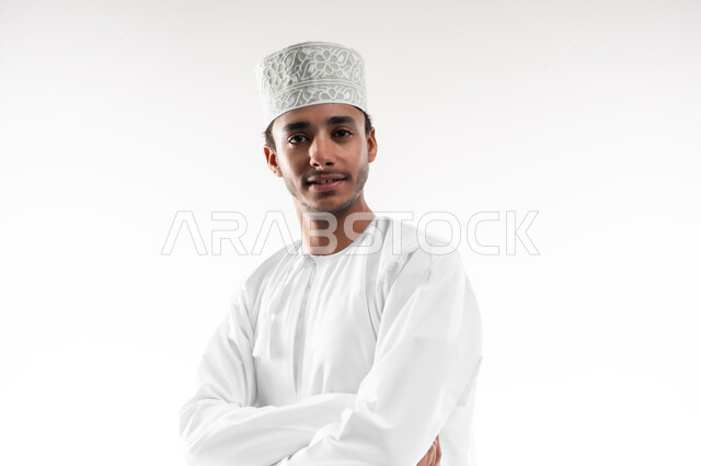 مفهوم الاناقة و الاهتمام بالمظهر الخارجي, بورتريه مقرب لشاب عربي خليجي عماني يرتدي الدشداشة و الكمه ينظر إلى الكاميرا بإيماءات السعادة و السرور، تعابير الرجولة والثقة بالنفس, خلفية بيضاء
