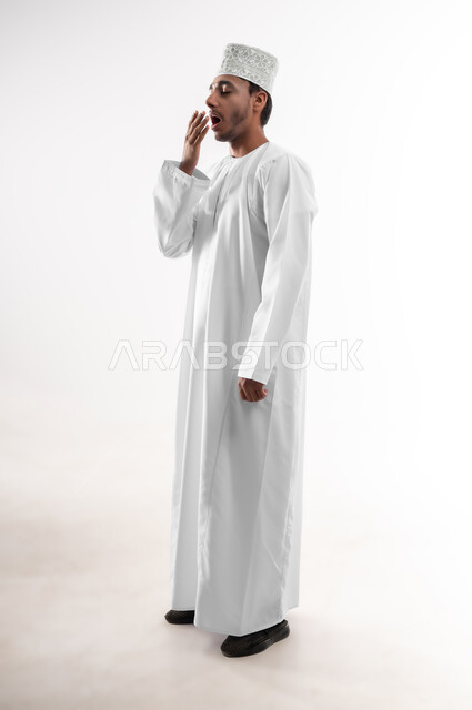 الإحساس بالكسل والخمول و الإرهاق، بورتريه لشاب عربي خليجي عماني يرتدي الدشداشة و الكمه يضع يده على فمه بإيماءات بإيماءات التثاؤب والنعاس، الشعور بالتعب والحاجة الى النوم والراحة, صوة الجسم بالطول الكامل، خلفية بيضاء