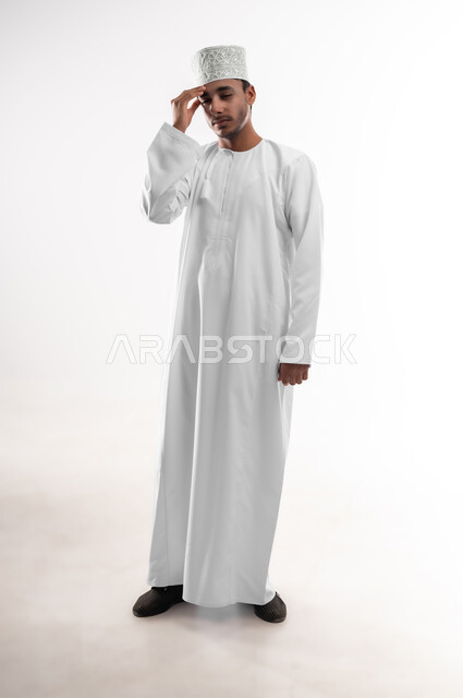 الحاجه للعلاج وطلب المساعده, بورتريه لشاب عربي خليجي عماني يرتدي الدشداشة و الكمه يضع يده على رأسه بإيماءات الدوار و الألم, الشعور بالتعب والإرهاق, الإصابة بنوبات الشقيقة، تعابير الصداع والإحساس بالمرض,صوة الجسم بالطول الكامل، خلفية بيضاء
