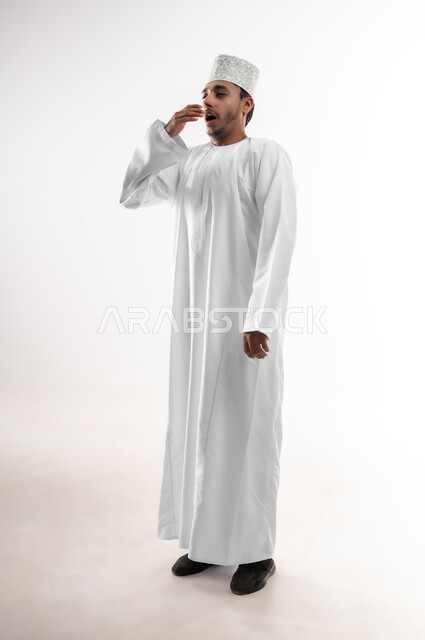 الشعور بالمرض و الزكام والحاجة الى العلاج والدواء, التهاب الأنف التحسسي, بورتريه لشاب عربي خليجي عماني يرتدي الدشداشة و الكمه يضع يديه على وجهه بإيماءات العطاس, صوة الجسم بالطول الكامل، خلفية بيضاء