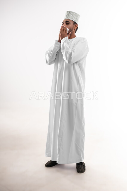 الشعور بالمرض و الزكام والحاجة الى العلاج والدواء, التهاب الأنف التحسسي, بورتريه لشاب عربي خليجي عماني يرتدي الدشداشة و الكمه يضع يديه على وجهه بإيماءات العطاس, استخدام المناديل الورقيه, صوة الجسم بالطول الكامل، خلفية بيضاء