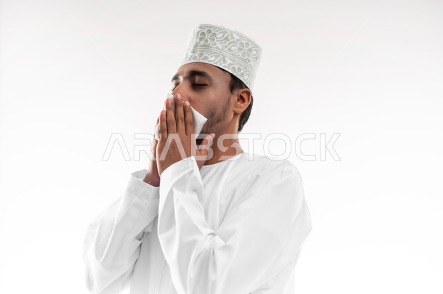 التهاب الأنف التحسسي, الشعور بالمرض و الزكام والحاجة الى العلاج والدواء, بورتريه مقرب لشاب عربي خليجي عماني يرتدي الدشداشة و الكمه يضع يديه على وجهه بإيماءات العطاس, استخدام المناديل الورقيه, خلفية بيضاء