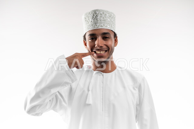 استخدام قشور فينير من الخزف لصنع ابتسامة بيضاء ناصعه، أهمية متابعة طبيب الاسنان، العناية والتجميل والتبيض، بورتريه مقرب لشاب عربي خليجي عماني يرتدي الدشداشة و الكمه ينظر الى الكاميرا يشير بيديه إلى أسنانه بإيماءات السرور، خلفية بيضاء