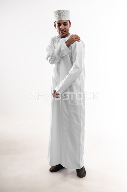 Muscle and joint inflammation, nerve aches and pains, request for help and need for treatment, portrait of a young Arab Gulf Omani man wearing a dishdasha and a cuff, putting his hand on his shoulder with gestures of pain, fatigue and exhaustion, emergency medical case, full body image, white background