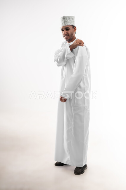 التهاب العضلات والمفاصل، الأوجاع والآلام العصبية، طلب المساعده والحاجه للعلاج، بورتريه لشاب عربي خليجي عماني يرتدي الدشداشة و الكمه يضع يده على كتفه بايماءات الألم والتعب والأرهاق، حالة اسعافية طارئة، صورة الجسم بالطول الكامل، خلفية بيضاء