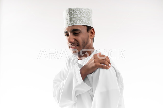 التهاب العضلات والمفاصل، الأوجاع والآلام العصبية، طلب المساعده والحاجه للعلاج، بورتريه مقرب لشاب عربي خليجي عماني يرتدي الدشداشة و الكمه يضع يده على كتفه بايماءات الألم والتعب والأرهاق، حالة اسعافية طارئة، خلفية بيضاء